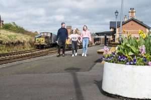 Father and daughters skipping along beside Whitehead railway museum with train in background and flower bed in the front